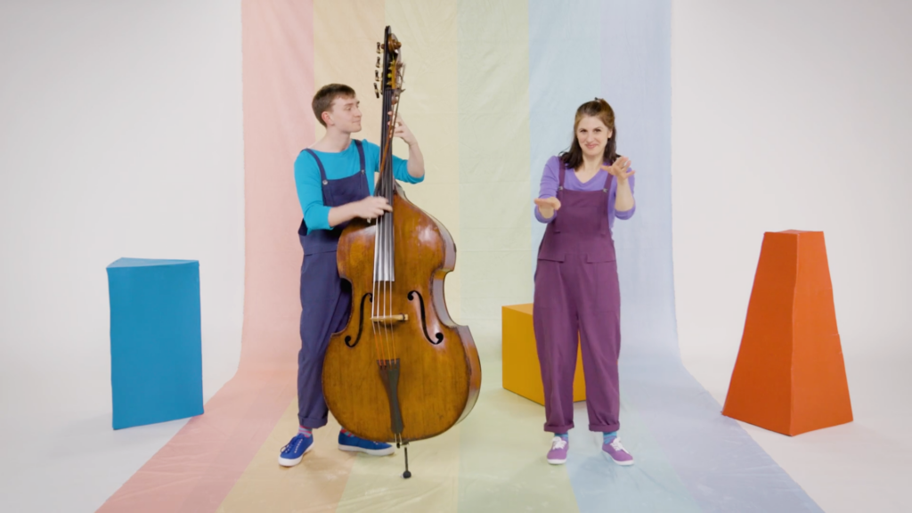 Two people stand in front of a rainbow that falls behind them to underneath their feet all the way to the front of the photo. There is a man playing double bass, and a woman doing the jive with her hands. Around and behind them are three coloured blocks - blue, yellow and orange